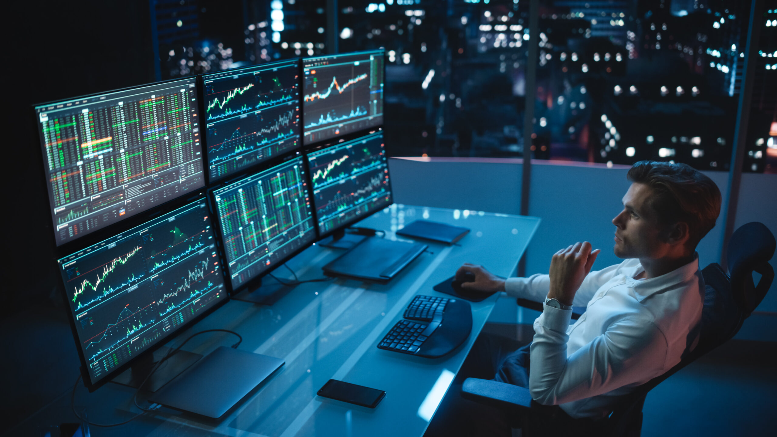 Financial Analyst Working on a Computer with Multi-Monitor Workstation with Real-Time Stocks, Commodities and Foreign Exchange Charts. Businessman Works in Investment Bank City Office Late Evening.